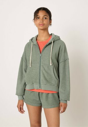 Felpa verde in spugna con zip frontale, grandi tasche e una texture morbida, abbinata a pantaloni corti coordinati. Fodera interna in arancione a contrasto.