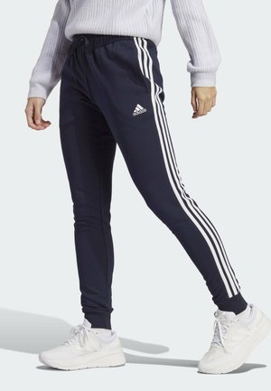 Personne portant un pantalon de survêtement Adidas bleu marine avec des bandes blanches, des baskets blanches et un pull en maille gris clair, debout dans une posture de marche.