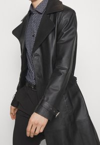 Zwart leren trenchcoat met brede revers en een getailleerde pasvorm, voorzien van knoopsluitingen en verstelbare manchetten. Draagt men over een patroon overhemd.