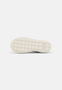 Semelle blanche de basket avec un motif en grille de croix en relief et un petit logo rectangulaire près du talon, présentée à plat sur un fond gris.