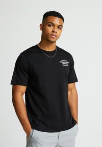 Must musta T-särk lühikeste varrukatega, millel on väike valge logo rinnal. Sirge äär ja ümar kaelus. Kantud heledate hallide pükste abil.