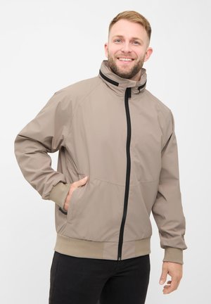 Smilende mand med kort hår iført beige lynlåstrøje og sorte bukser, stående med den ene hånd i jakke lommen mod en hvid baggrund.