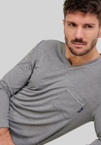 Camiseta gris de manga larga en tejido suave con cuello en V. Presenta un bolsillo en el pecho con ribete en contraste y una pequeña etiqueta de logo.