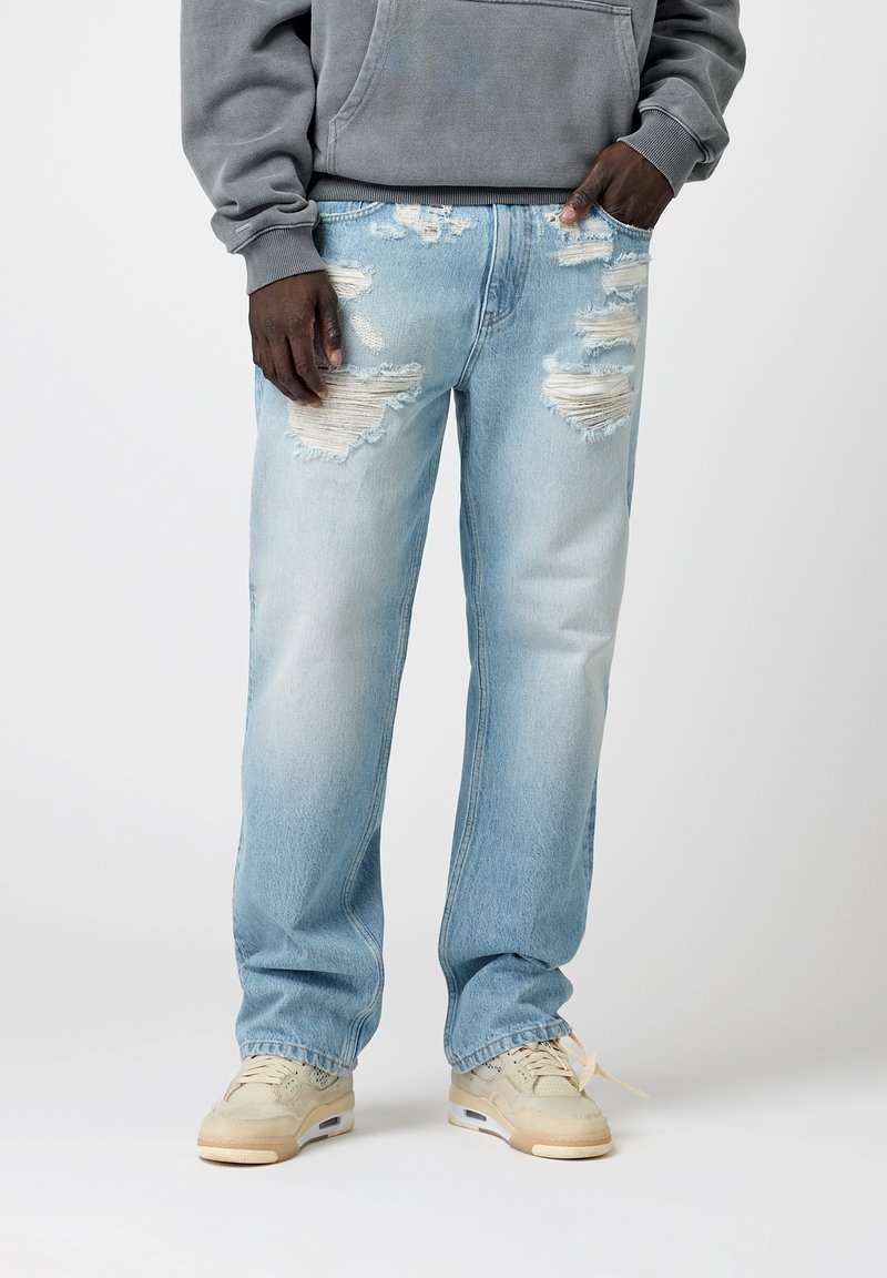 Jeans de mezclilla azul claro con patrones rasgados, que presentan un corte holgado y dobladillos deshilachados, combinados con zapatillas beige y una sudadera gris.