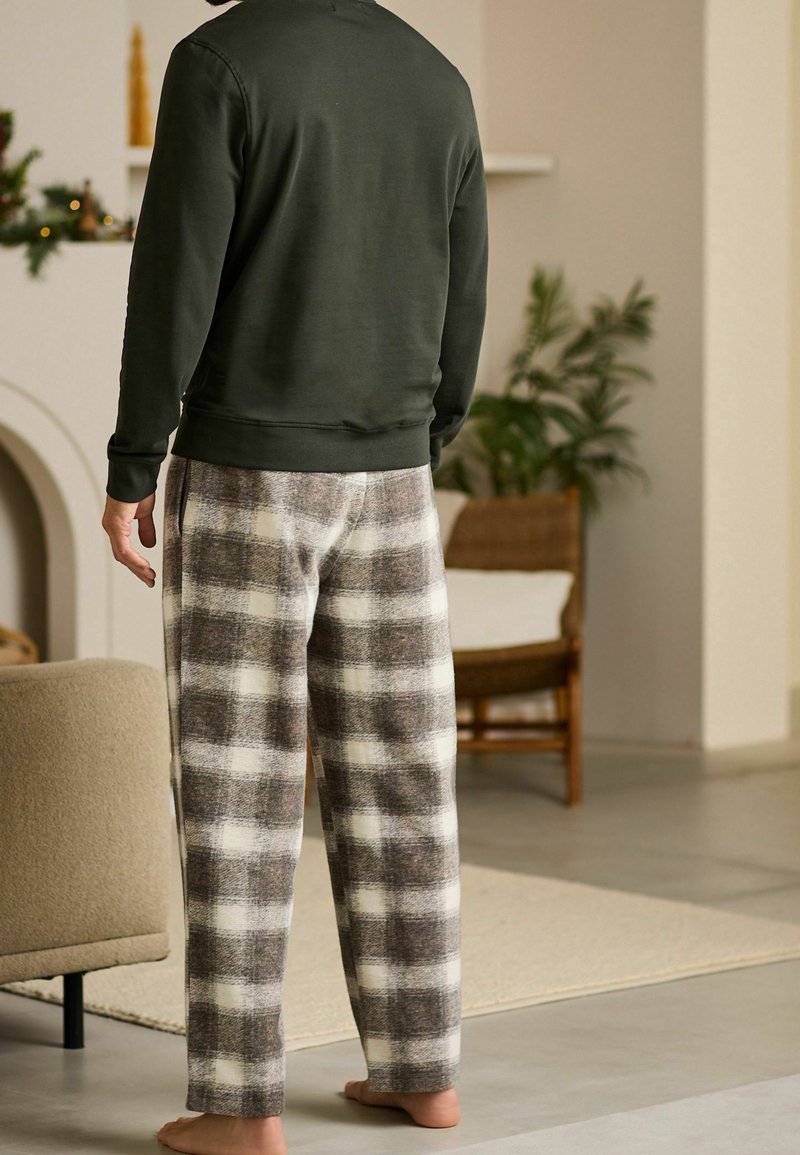 Homme debout pieds nus à l'intérieur, portant un pantalon de pyjama en polaire à carreaux et un haut sombre à manches longues avec un fond de salon chaleureux.