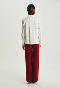 Blouse blanche à manches longues avec un pli au dos, associée à un pantalon large rouge taille haute avec des rayures latérales et une texture lisse.