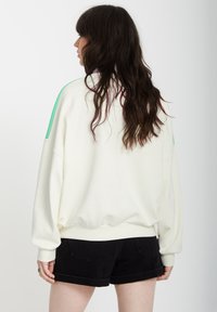 Weißes oversized Sweatshirt mit grünen Schulterstreifen, rundem Ausschnitt und gerippten Bündchen. Kombiniert mit schwarzen Shorts; glatte Textur.