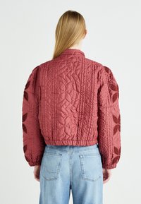 Prošívaná maroon bomber bunda s embosovaným květinovým vzorem, s úzkým pasem a nabíranými rukávy, kombinovaná se světle modrými džínami.