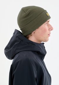 Olivgrüne gestrickte Mütze mit umgeschlagenem Bündchen, getragen mit einer marineblauen wasserdichten Jacke, die eine glatte Textur und ein minimalistisches Design zeigt.