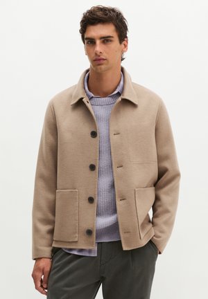 Young man wearing a beige buttoned jacket over a light purple sweater and dark pants, standing against a plain background.