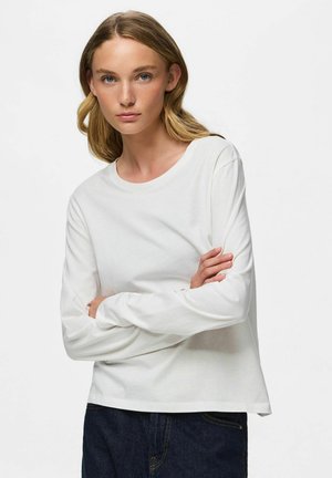 Jeune femme aux cheveux blonds ondulés, portant un t-shirt blanc uni à manches longues et un jean foncé, debout, les bras croisés devant un fond uni.