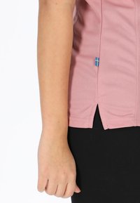 Camicetta corta a maniche corte di colore rosa chiaro in tessuto traspirante, con spacchi laterali, dotata di un'etichetta di accentuazione blu e gialla all'orlo.