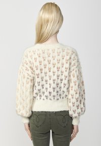 Pulover tricotat în ivory cu un model texturat de iepurași pastel. Are mâneci largi și un tiv ribatat. Este purtat cu pantaloni de culoare verde măsliniu.
