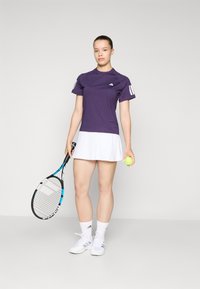 Paarse sportshirt met korte mouwen, witte rok, houdend een tennisracket en bal, witte sneakers en enkelsokken.
