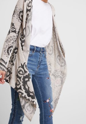 Person trägt blaue bestickte Jeans, weißes T-Shirt und einen beigen offenen Cardigan mit schwarzen, kunstvollen Blumenmustern.