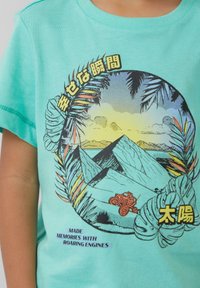 Turquoise katoenen T-shirt met een grafisch ontwerp van bergen, een voertuig en tropische bladeren. De tekst bevat meerstalige zinnen.