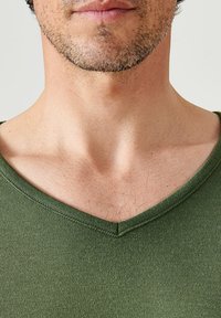 Groene v-hals t-shirt van een zachte, gladde stof. Close-up toont de halslijn, stikseldetails en lichte borsthaar tegen een egale huidskleur.