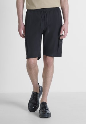 Bermudas negras de carga hechas de tela ligera, con cinturilla elástica, bolsillos laterales y un corte holgado. Combinadas con zapatos negros.