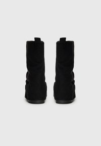 Botas de tornozelo em camurça preta com um design afundado, sola plana e alças de puxar na parte de trás. Textura suave sem padrões visíveis.