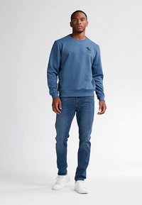 Modrá mikina z měkké, texturované látky s kulatým výstřihem a malým logem na hrudi, doplněná úzkými modrými džíny a bílými teniskami.