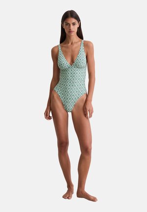 Femme debout pieds nus portant un maillot de bain une pièce à motif vert et blanc avec un décolleté en V profond et des bretelles fines.