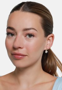 Boucles d'oreilles en or en forme de cercle avec un détail de pierre précieuse transparente et ronde. Texture lisse et design délicat, portées par une personne avec un maquillage neutre.