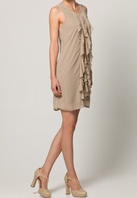 Beżowa sukienka bez rękawów z centralnym detalem falbany, gładką fakturą i prostym krojem. Noszona z beżowymi szpilkami.