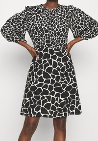 Svart och vit giraffmönstrad klänning med puffärmar, rund halsringning och figursydd överdel som övergår till en utsvängd kjol. Slät textur.