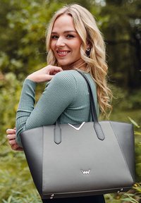 Graue Tote-Tasche mit strukturierter Form, ausgestattet mit schwarzen Griffen und einem silbernen Akzent. Glatte Textur mit einem subtilen Logo auf der Vorderseite.