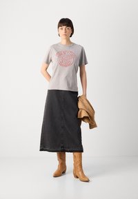 Graues Baumwoll-T-Shirt mit rotfarbigem Grafikdruck, kombiniert mit einem schwarzen Denim-Maxikleid und braunem knielangem Stiefeln, während sie eine braune Jacke hält.