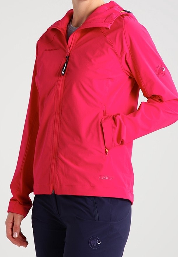 Personne portant une veste softshell rose vif avec des poches zippées et un pantalon foncé, debout avec une main dans la poche devant un fond uni.