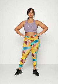 Sutiã desportivo roxo com um logo da Nike, combinado com leggings multicoloridas que apresentam padrões geométricos em amarelo, verde-água e rosa. Ténis de desporto pretos.