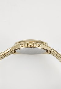 Vista laterale di un orologio da polso d'oro che mostra la sua corona e due pulsanti su un bracciale metallico a maglie su uno sfondo bianco.