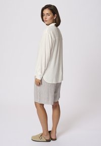 Witte blouse met lange mouwen en een ontspannen pasvorm, gecombineerd met gestreepte beige shorts. Het model draagt lichte tan klompen. Eenvoudig ontwerp, zachte textuur.