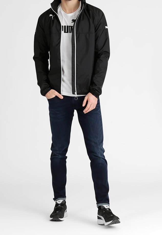Homme portant une veste noire zippée, un t-shirt blanc Puma, un jean foncé et des baskets noires Puma, debout les mains dans les poches.