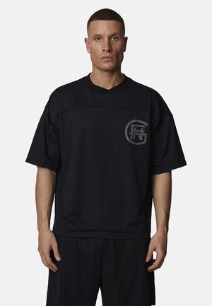 Homme debout portant une chemise en mesh noir à manches courtes ample et un short assorti avec un logo circulaire gris clair sur la poitrine.