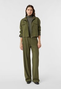 Oliwkowa skrócona kurtka z dwoma kieszeniami na piersi, nałożona na paskowany top z wysokim kołnierzem, zestawiona z szerokimi spodniami w tym samym kolorze. Czarne buty.