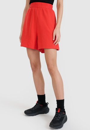 Osoba ubrana w jasnoczerwone luźne szorty, czarne skarpetki i czarne buty sportowe stojąca na jednolitym, jasnym tle.