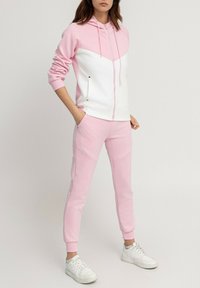 Haut à capuche rose et blanc avec fermeture éclair, présentant un design en chevron. Accompagné d'un pantalon rose, le tout en tissu doux. Baskets blanches.