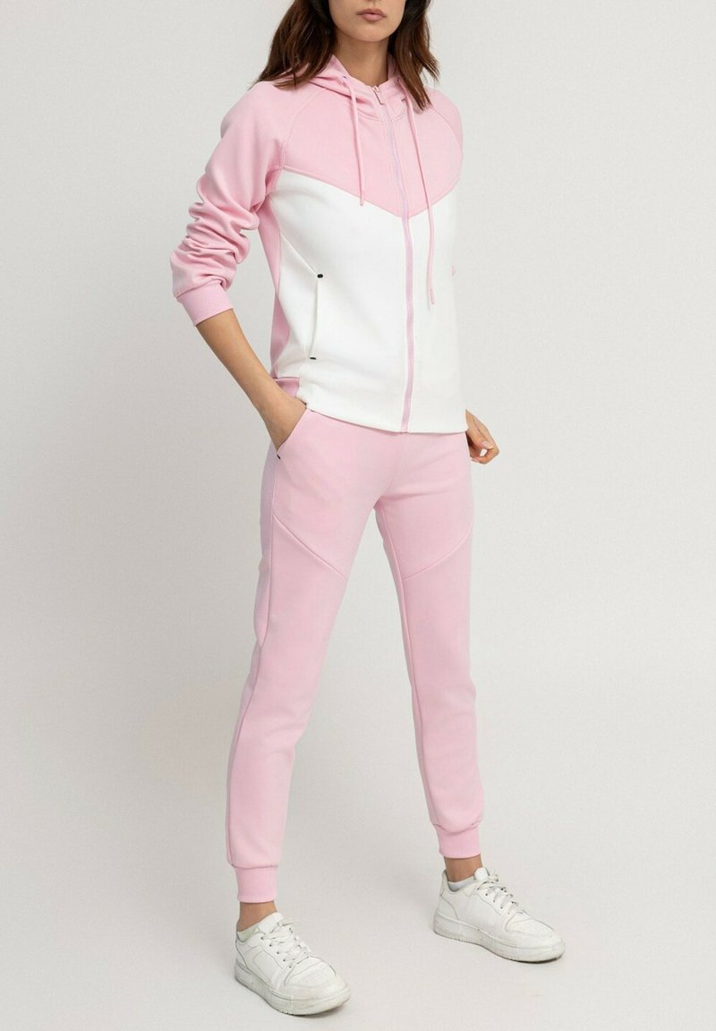 Haut à capuche rose et blanc avec fermeture éclair, présentant un design en chevron. Accompagné d'un pantalon rose, le tout en tissu doux. Baskets blanches.