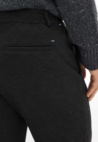 Close-up van een persoon die donkere, getextureerde broek en een grijze gebreide trui draagt, met zicht op een achterzak en een klein logolabel.