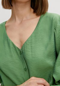 Blouse verte à manches courtes avec un col en V et fermeture à bouton. Le tissu a une surface lisse et légèrement texturée. Design doux et léger.