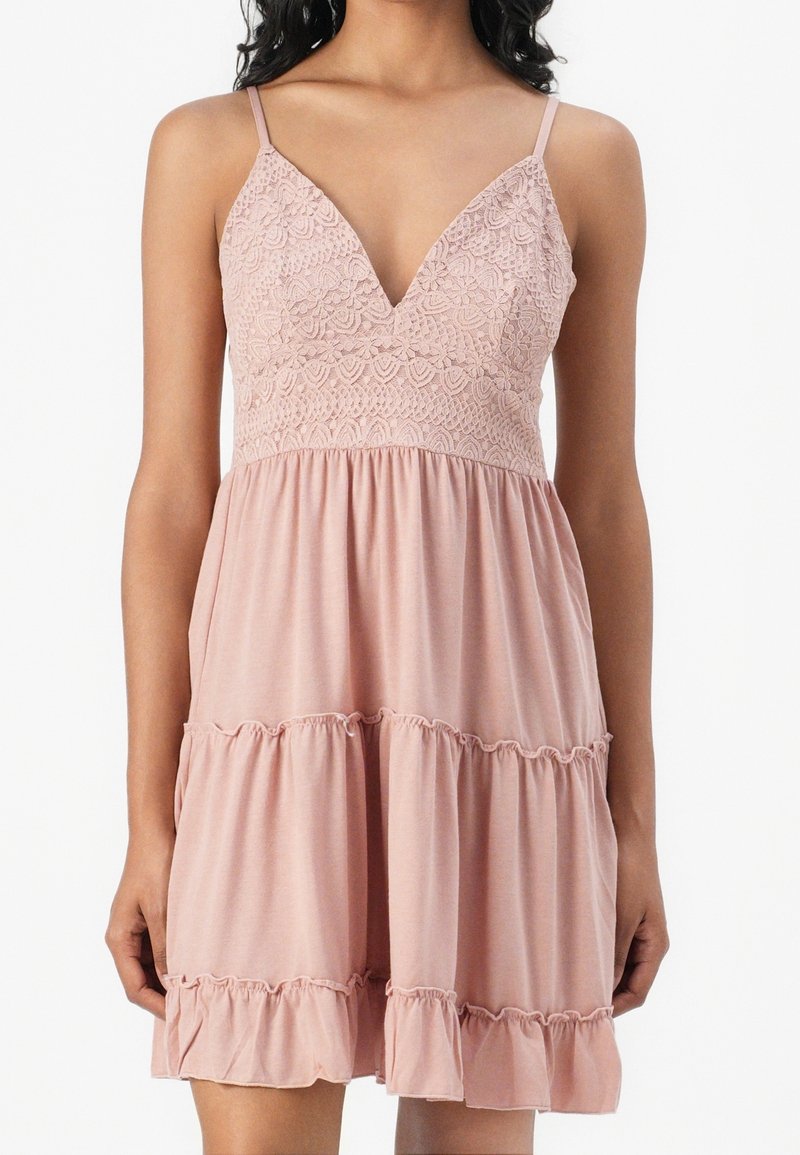 Femme portant une robe rose pâle sans manches avec un corsage en dentelle, fines bretelles, jupe à étages et détails à volants à l’ourlet.