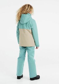 Waterdichte jas in mintgroen en beige, met een capuchon en elastische manchetten, gecombineerd met bijpassende mintkleurige broek en zwarte laarzen.