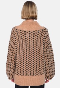 Person med kort blont hår bär en oversized tanfärgad tröja med svart geometriskt mönster, ribbad krage, muddar och fåll, vänd bort från kameran.