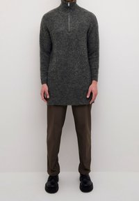 Grauer, gestrickter Pullover mit langen Ärmeln und Viertelreißverschluss, kombiniert mit braunen Hosen und schwarzen Plateau-Schuhen. Glatte Textur überall.