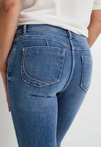 Jeans de denim azul con un diseño ajustado, cintura de altura media y dos bolsillos traseros con costura curvada. El tejido tiene una textura suave.