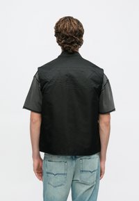 Schwarze ärmellose Jacke mit strukturiertem Rücken und abgerundetem Design, kombiniert mit einem grauen Kurzarmhemd und hellblauen Jeans.