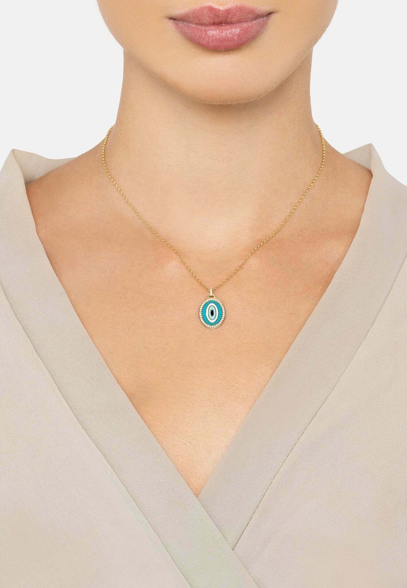 Kultaketju, jossa on soikea turkoosi riipus mustilla ja valkoisilla yksityiskohdilla, vaaleanharmaata vaatetta vasten.