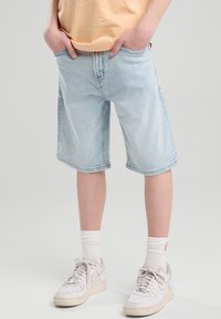 Un bambino indossa una maglietta arancione e pantaloni corti in jeans blu chiaro, con le mani nelle tasche, abbinati a sneakers bianche e calzini, su uno sfondo neutro.